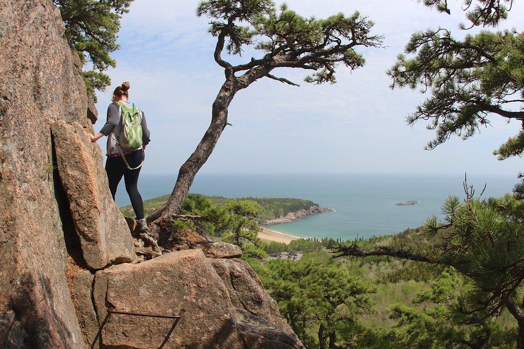 A hiker navigates a rocky cliffside along the Beehive Trail overlooking Sand Beach in Acadia National Park.