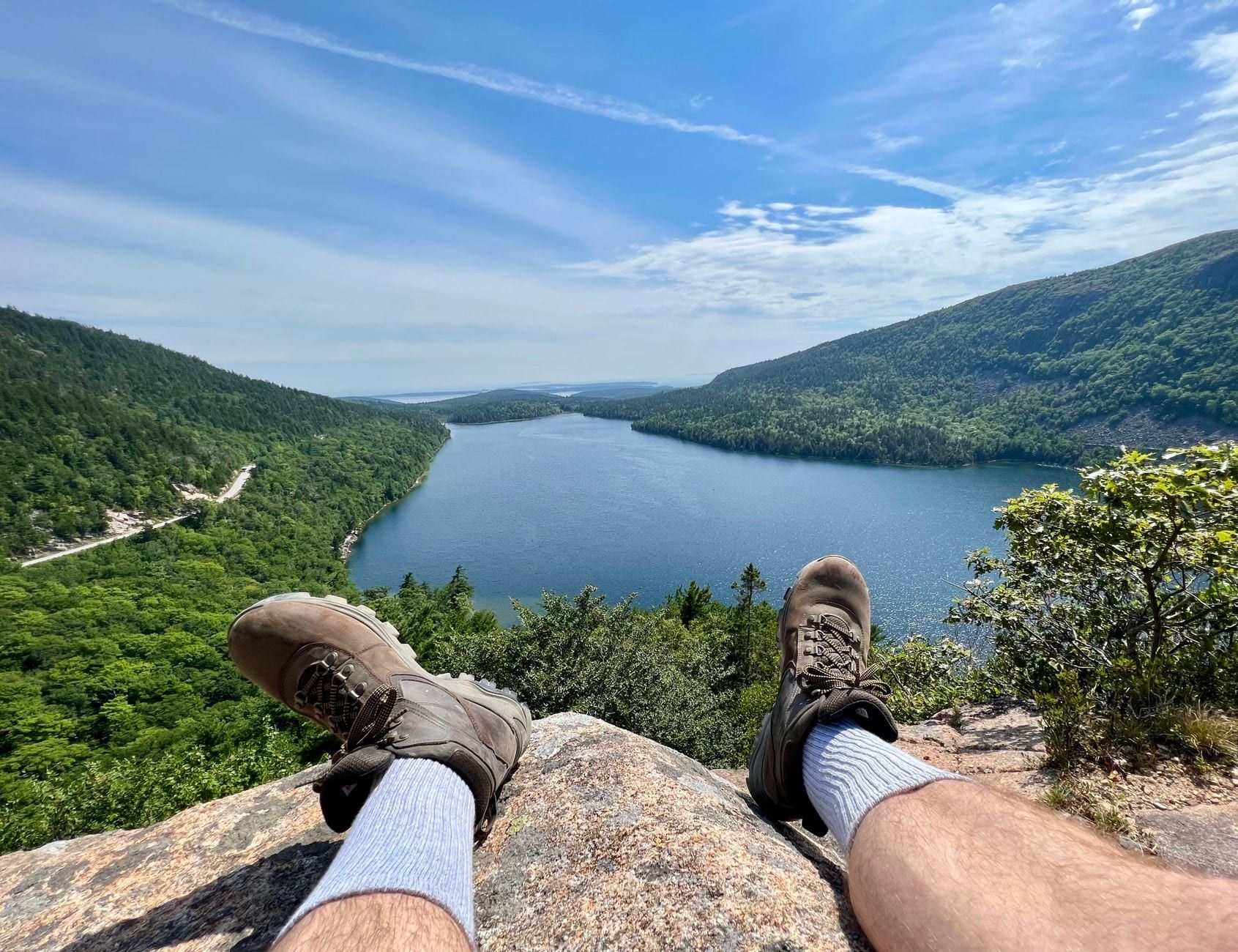 Die Wanderschuhe eines Wanderers auf einem felsigen Vorsprung mit Blick auf das Wasser und die bewaldeten Hügel des Jordan Pond im Acadia-Nationalpark.