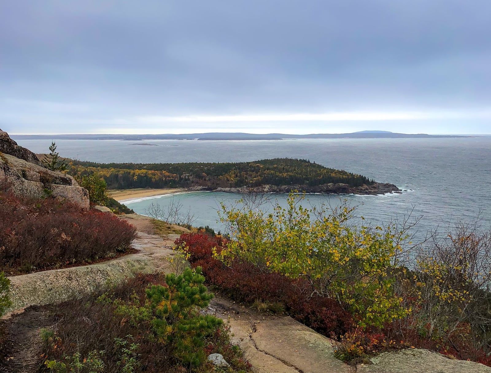 Herbstlaub säumt einen Granit-Wanderweg mit Blick auf den Sand Beach und den Atlantischen Ozean im Acadia-Nationalpark.