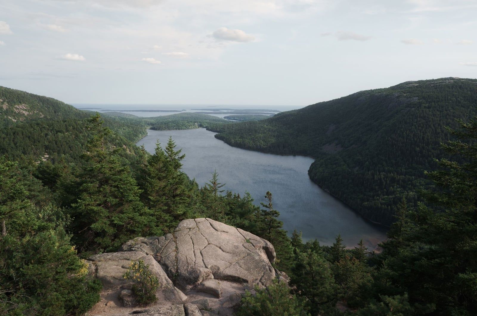 Ein von immergrünen Bäumen eingerahmter, felsiger Gipfelaussichtspunkt blickt im Acadia-Nationalpark auf den Jordan Pond und die fernen Ozeaninseln herab.