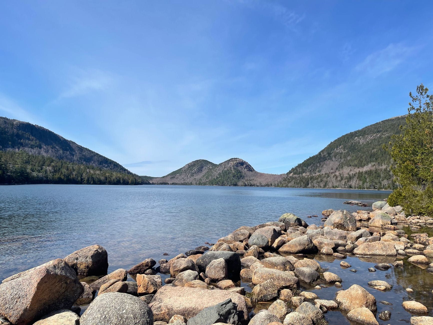 A rocky shoreline borders Jordan Pond, looking across clear water toward the North and South Bubbles in Acadia National Park under a bright blue sky.