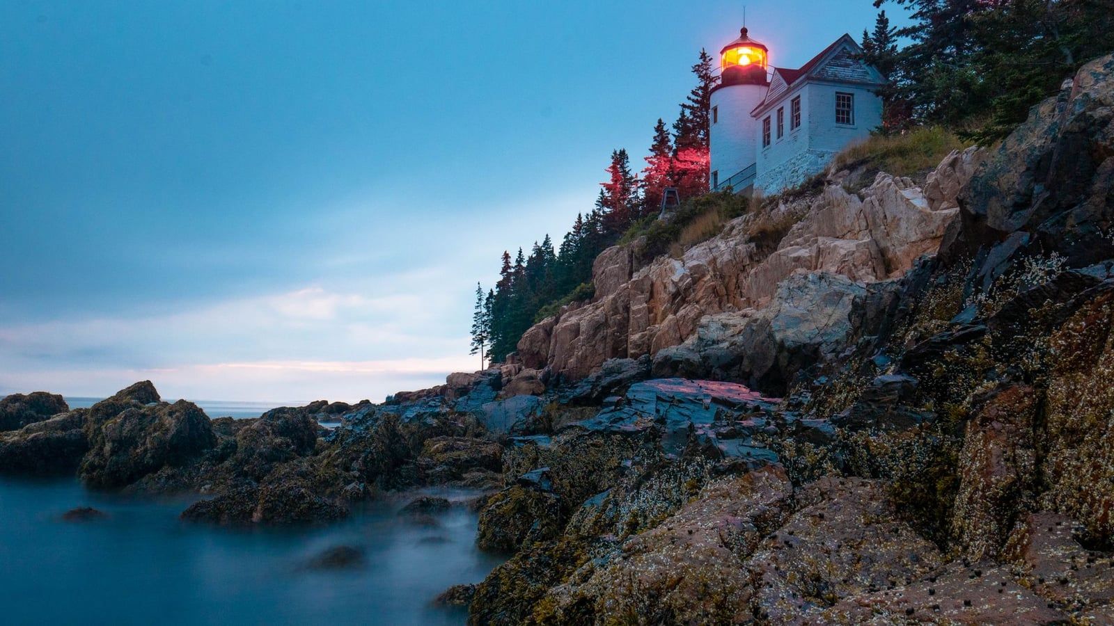 Bass Harbor Head Lighthouse shines with a red beacon atop rocky ocean cliffs at twilight in Acadia National Park.